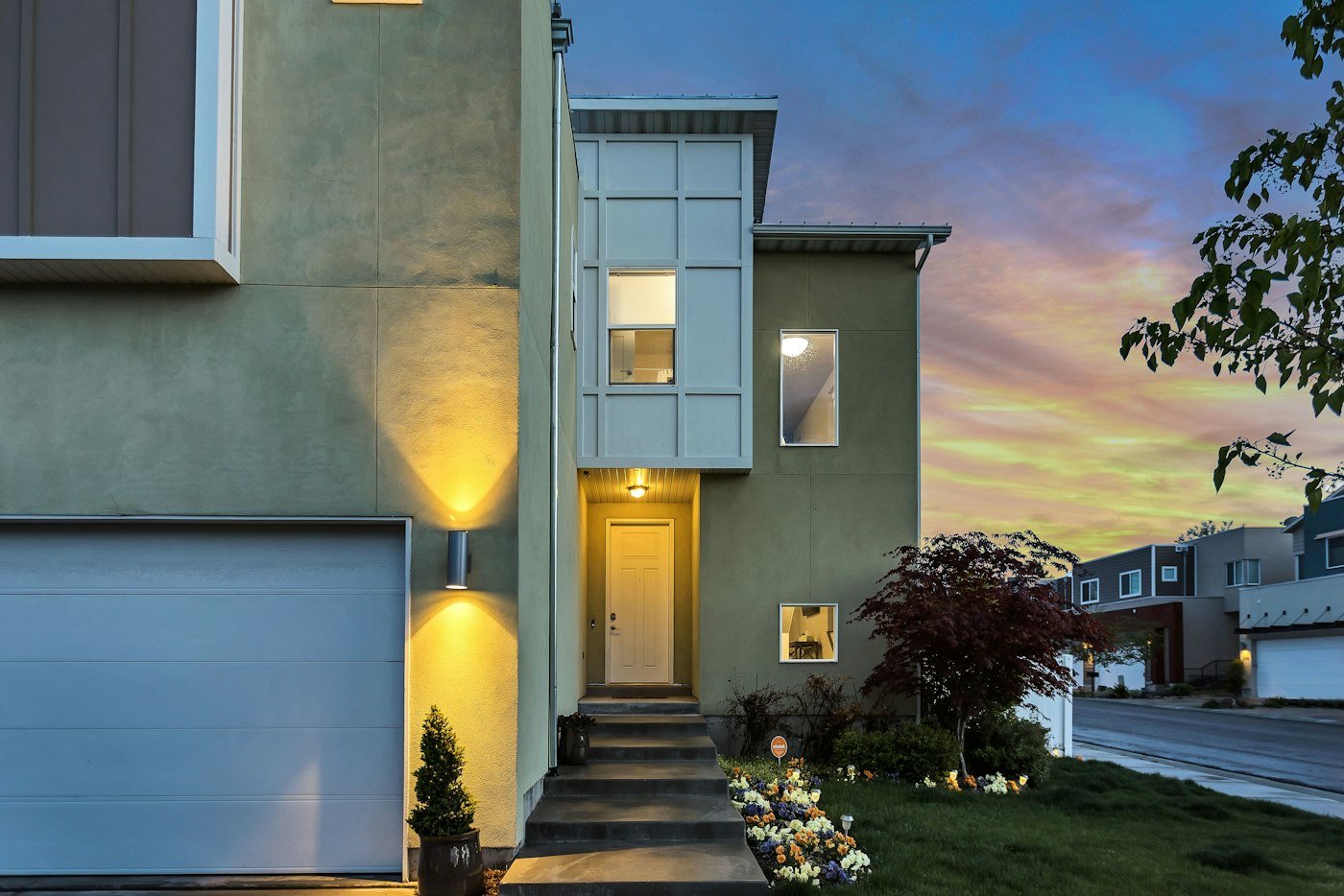 A sage green house at dusk with a white front door, a pale garage door, and steps down to a flower bed